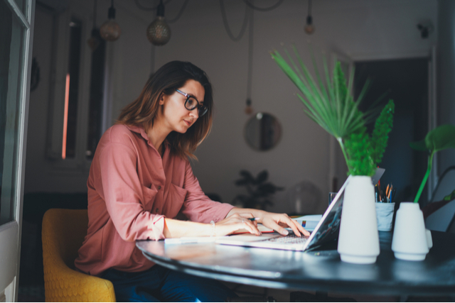 side view of woman sat at table with laptop, pen in hand, focussing on screen