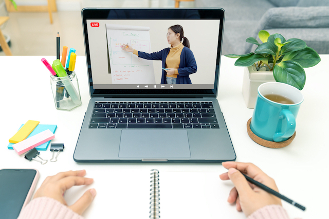 point of voiew of an attendee at an online class. Laptop with tacher on screen, student's hands and notebook